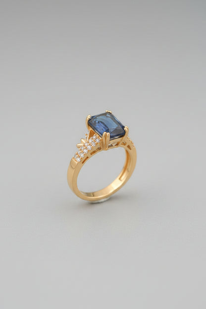 Gold ring with a blue gemstone on a reflective surface, blurred background