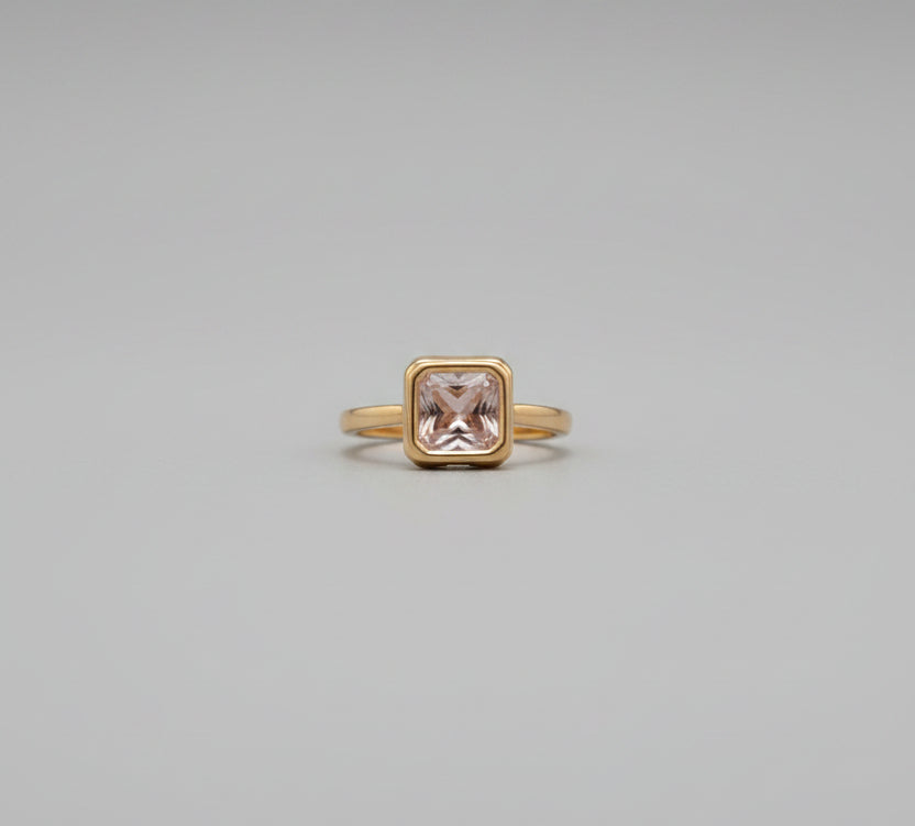 Gold ring with a pink gemstone on a reflective surface, blurred background